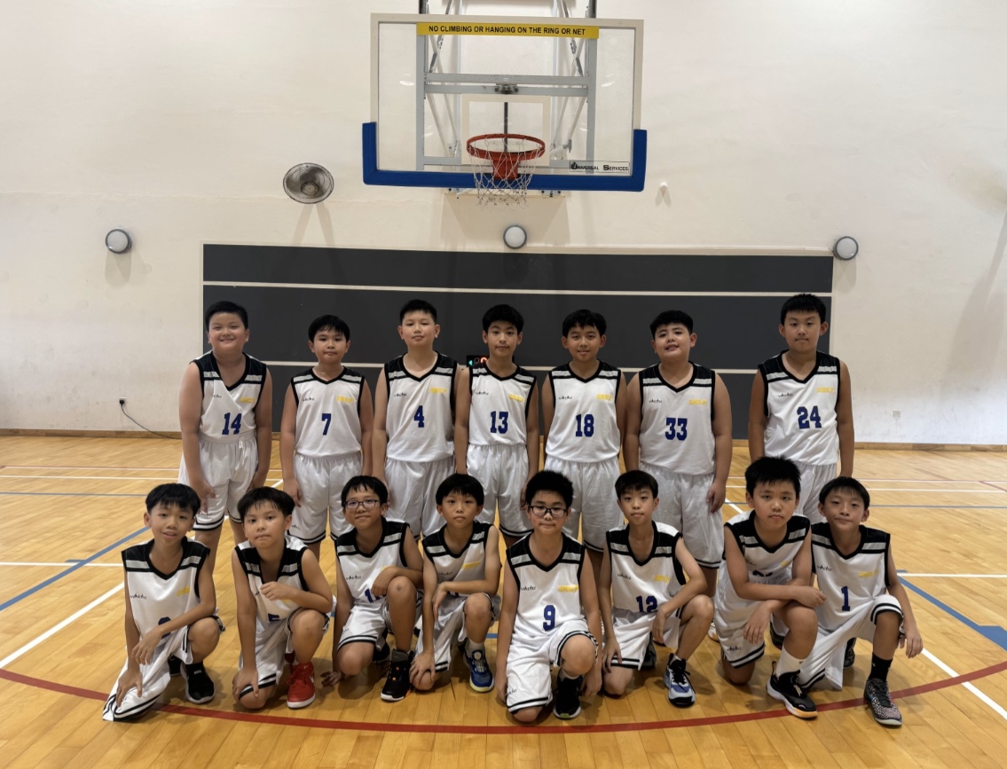 Photo 8: A team of 15 primary school boys in matching white and black basketball jerseys pose for a team photo inside a school gymnasium, arranged in two rows — seven standing at the back and eight kneeling in front — in front of a basketball hoop.